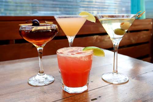 Four cocktails arranged on a wooden table — an amber coupe drink garnished with blueberries, a pale orange martini, a pink-red iced drink with a salted rim and lime wedge, and a classic martini garnished with green olives.