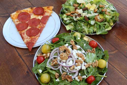 Three menu items on a wooden table: a slice of pepperoni pizza on a white plate, a Caesar salad with croutons and shaved parmesan, and a spinach salad with cherry tomatoes, red onion, walnuts, and crumbled feta.