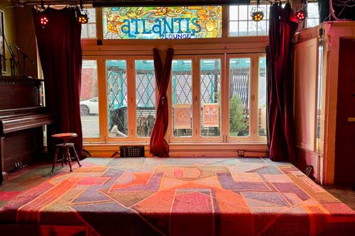 The stage at Atlantis Lounge, featuring a colorful patterned carpet, a piano to the left, red stage curtains, and a decorative stained glass window with the "Atlantis Lounge" name above large windows overlooking the street.
